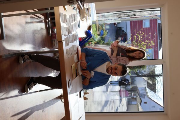 1 Frau steht im Hintergrund, ein Mann sitzt am Anmeldetisch, beide blicken in die Kamera