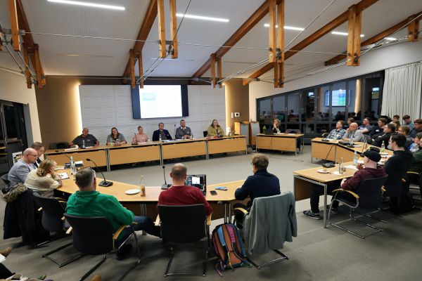 circa 25 Personen im Kreis sitzen, mit Tischen